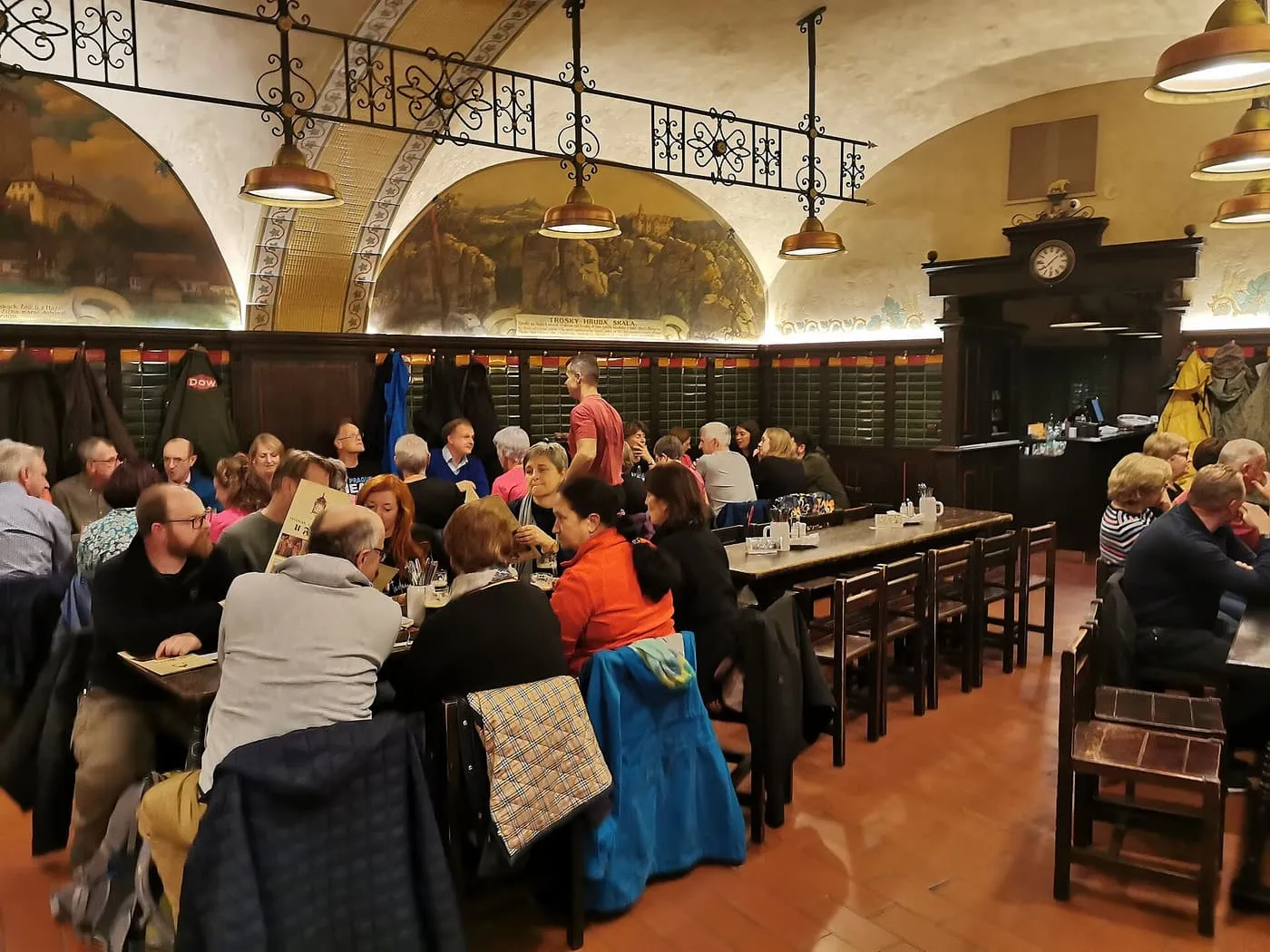 A vaulted beer hall at U Fleků with long wooden tables, painted lunettes on the walls and diners seated at the tables