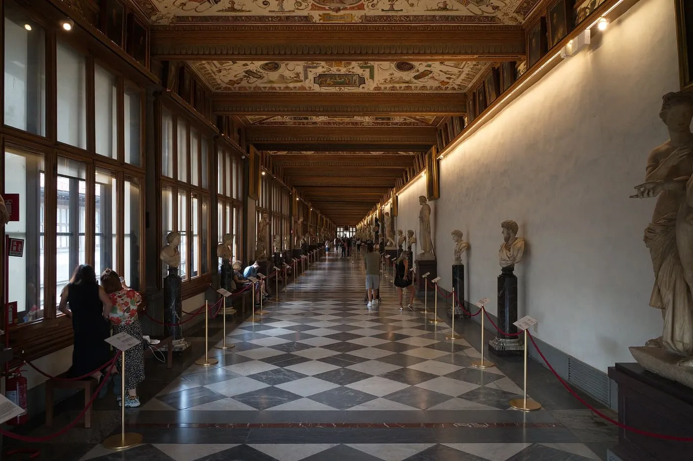 A long Uffizi corridor with a chequered marble floor, ancient busts along the windows and a painted ceiling