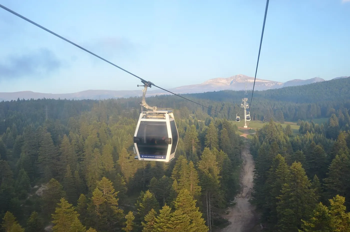 A cable car cabin hanging from its cable above a forest of conifers on the way up Uludağ, with a bare mountain beyond