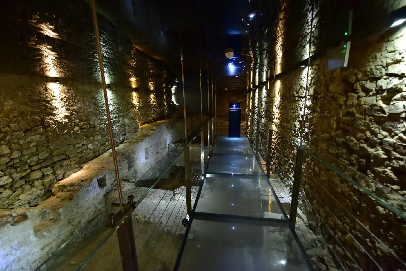 A glass walkway between stone walls of the excavations under the Main Market Square, lit by spotlights