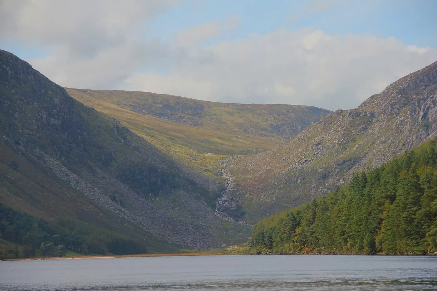 The Upper Lake at Glendalough with steep glacial valley sides, a thin waterfall and conifer forest on the right