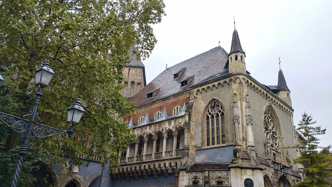 The Gothic wing of Vajdahunyad Castle with a pointed tower behind trees under a grey sky