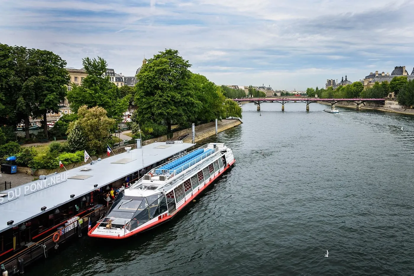 A red-and-white Vedettes du Pont Neuf boat moored at its pontoon beside the tree-lined tip of the Île de la Cité, with the Pont des Arts in the distance