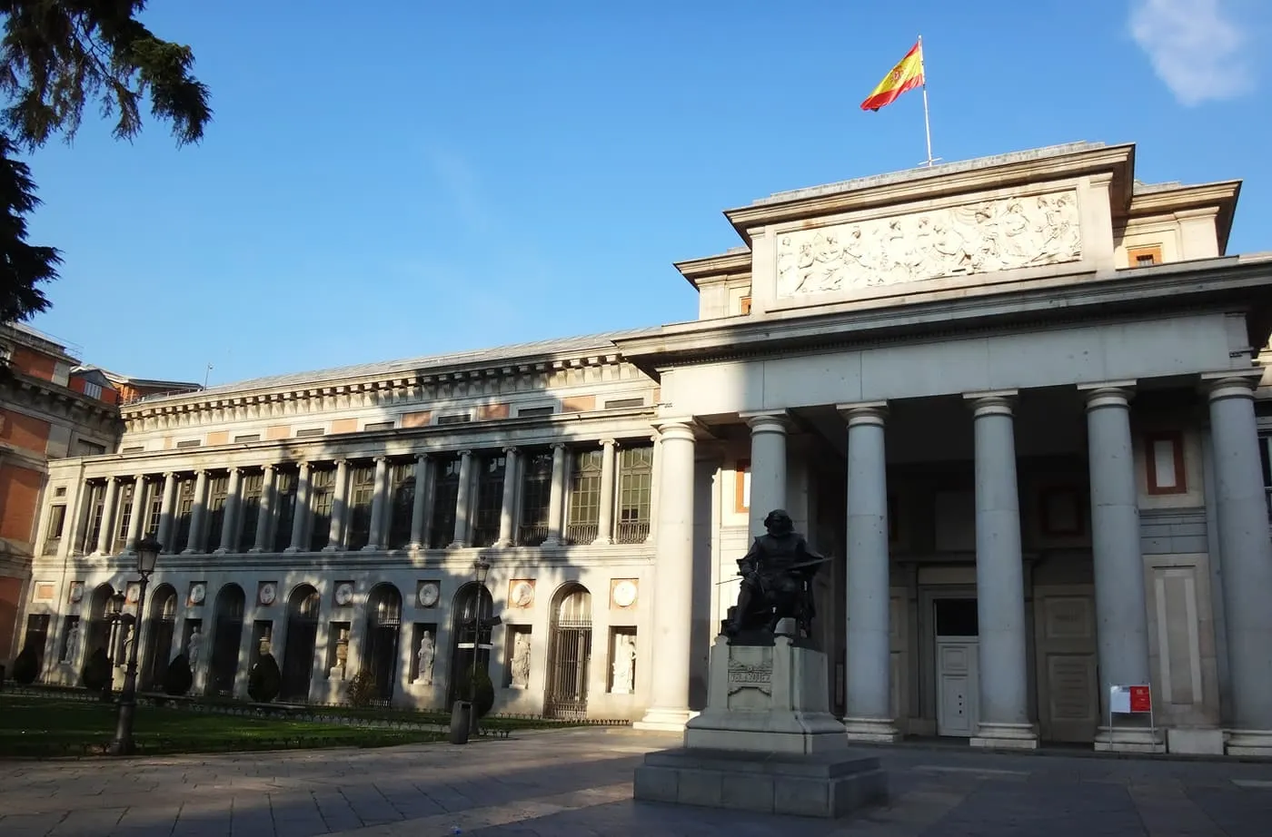 The columned Velázquez door of the Prado Museum, with the bronze statue of Velázquez in front and a Spanish flag above