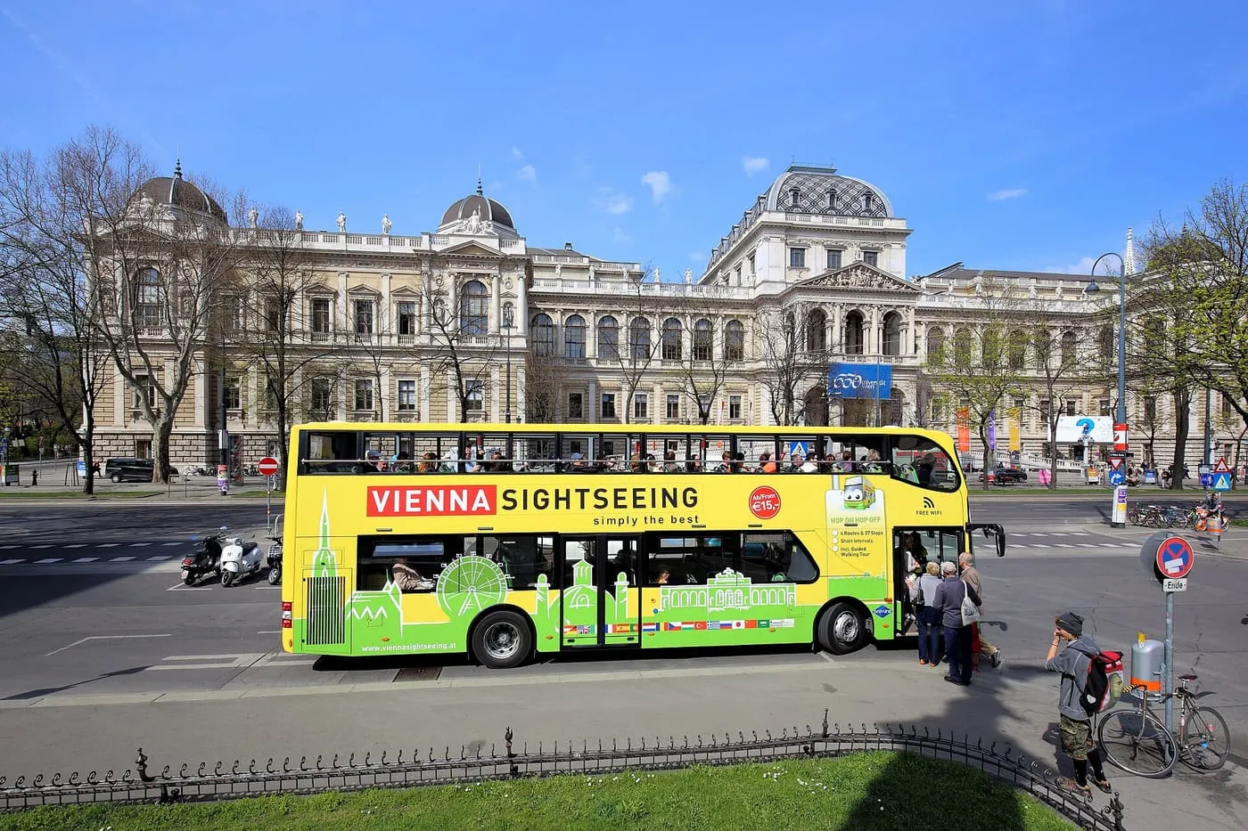 A yellow Vienna Sightseeing double-decker bus at a stop on the Ring, with the main university building behind