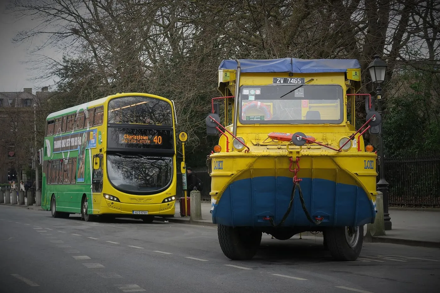 A yellow and blue amphibious DUKW seen head-on at the kerb, with a Dublin Bus double-decker at a stop behind it