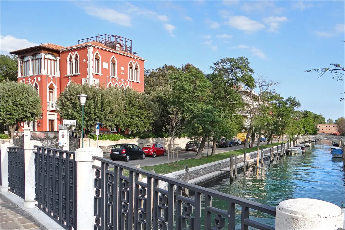 A red Gothic-style villa behind trees beside a canal with a paved embankment on the Lido