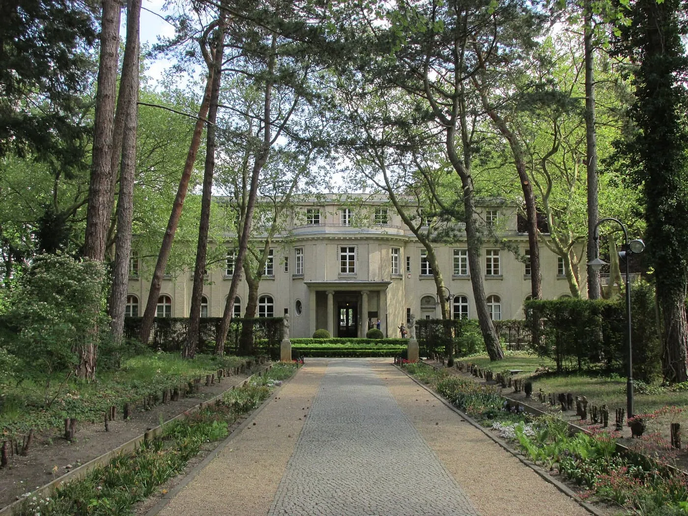 The House of the Wannsee Conference, a pale villa seen along a gravel and cobbled garden path between tall pines