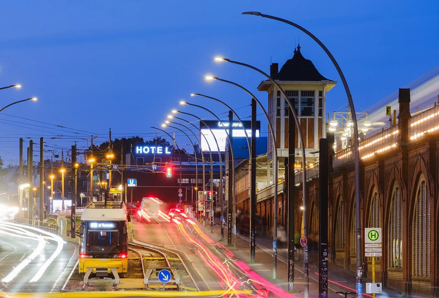 A yellow tram and trails of car lights on Warschauer Straße at dusk, with the brick arches of the elevated U-Bahn line on the right