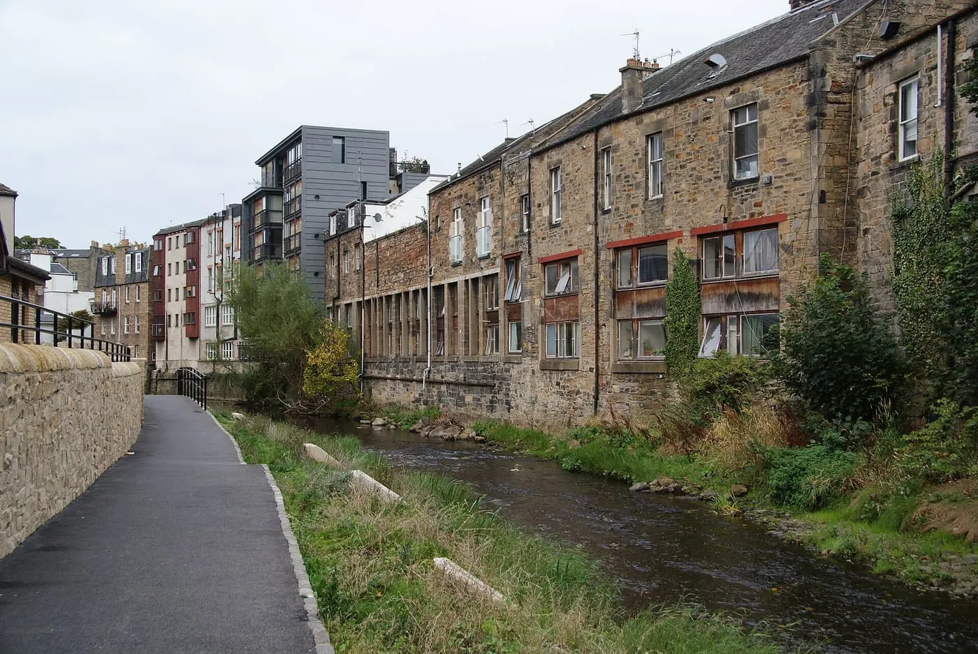 The Water of Leith running between a grassy bank and old stone buildings, with a tarmac path alongside, at Stockbridge