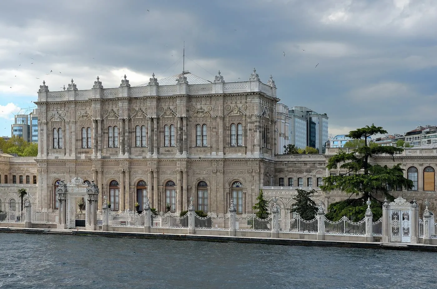 The long stone facade of Dolmabahçe Palace behind a white iron fence on the Bosphorus shore, under a cloudy sky