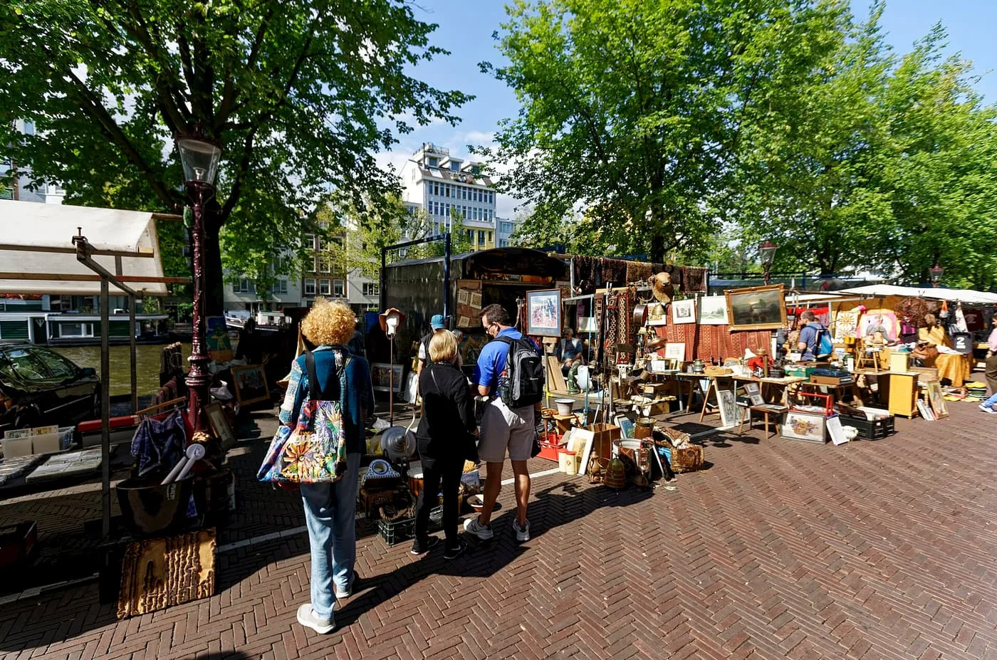 Shoppers looking at antiques and second-hand goods on a market stall under trees beside a canal