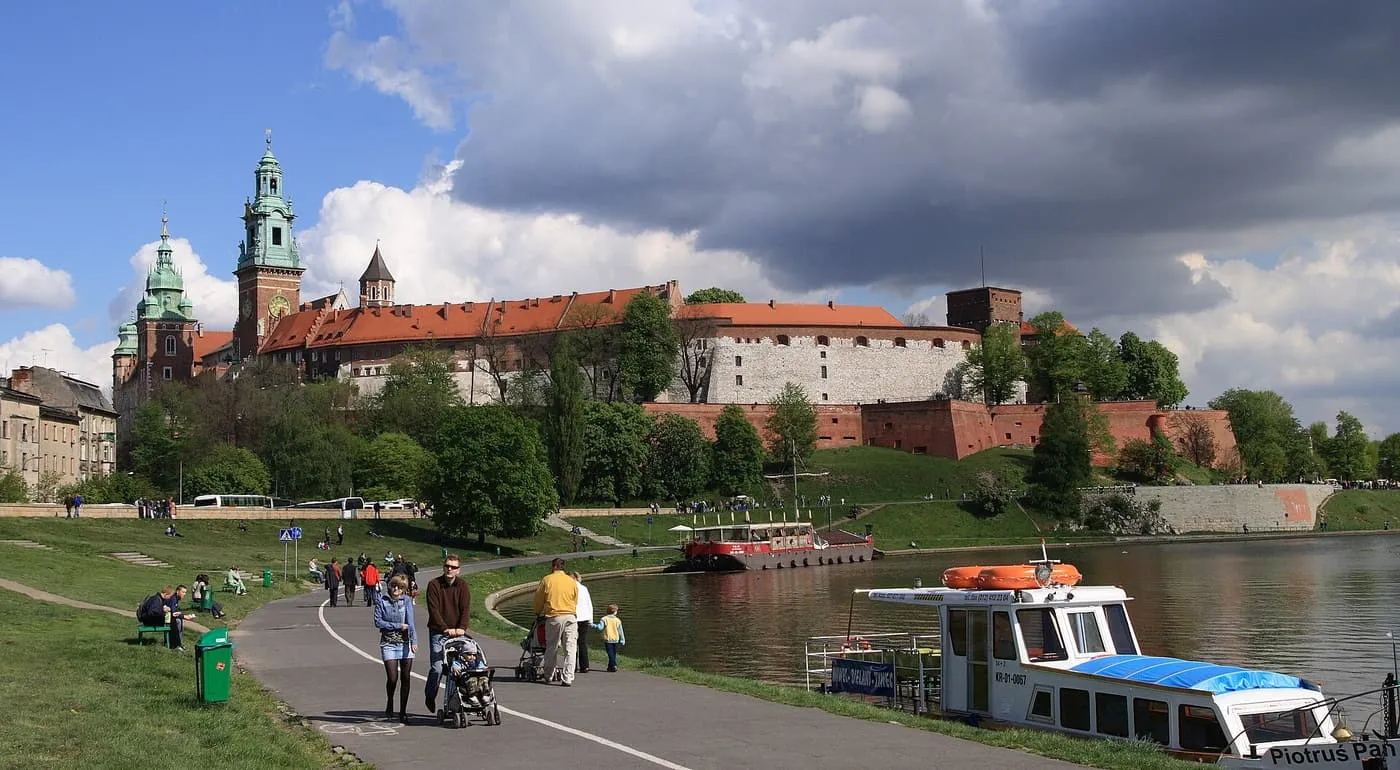 Wawel Hill and its castle walls above the Vistula, seen from the riverside path with walkers and moored boats
