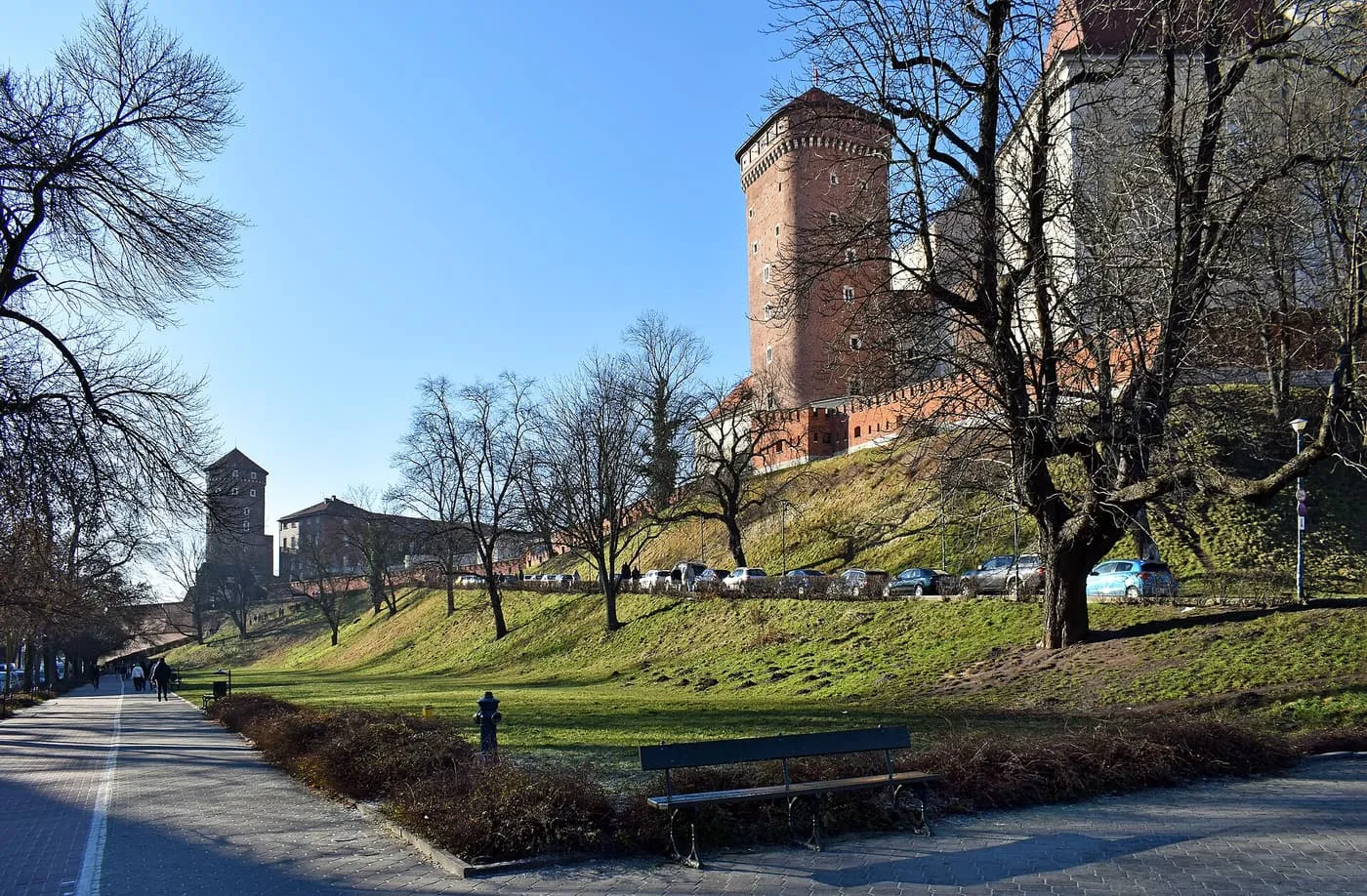 The red-brick walls and tower of the Wawel above a grassy slope and a paved path, under a clear blue sky