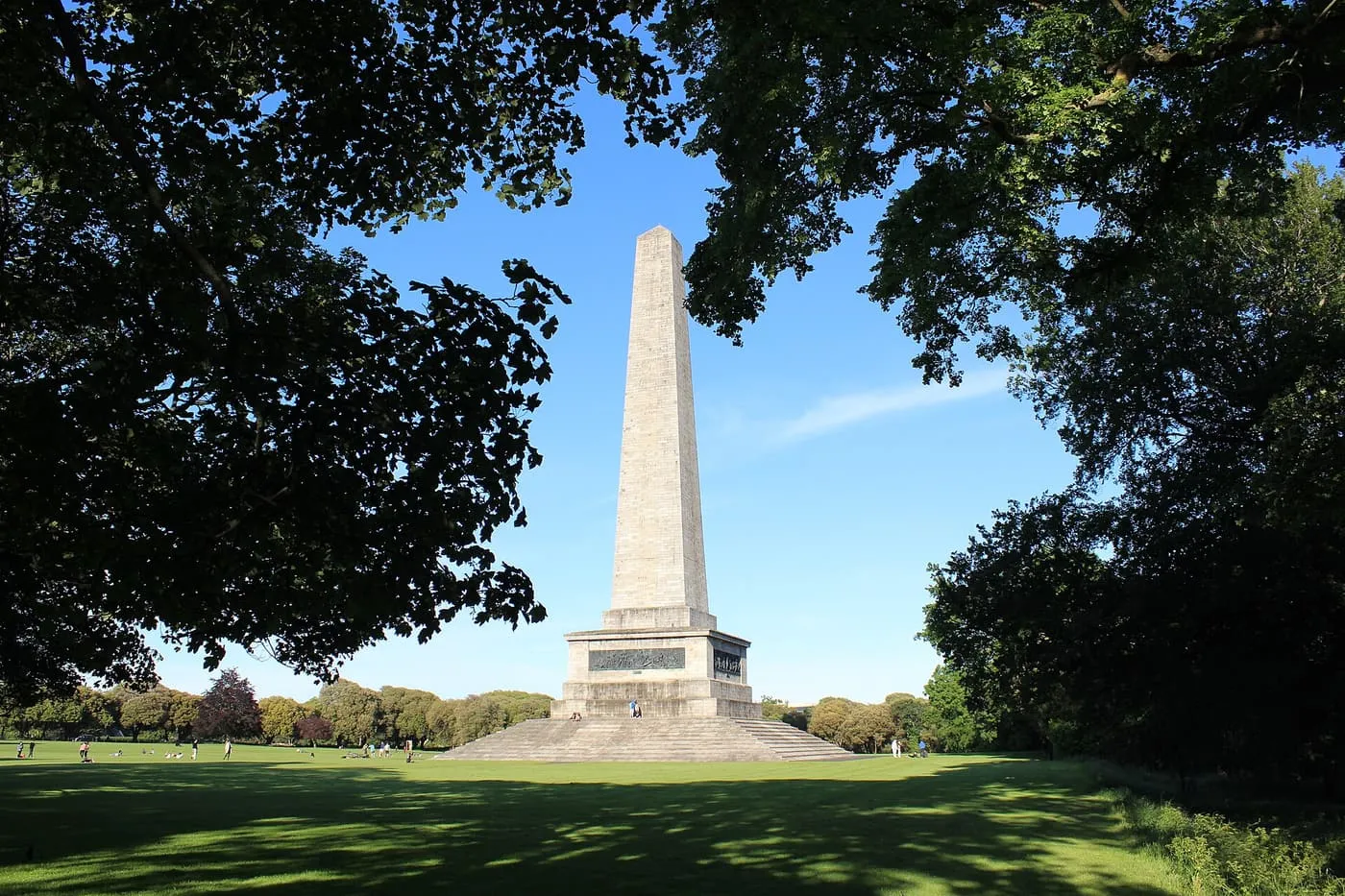 The Wellington Testimonial obelisk seen through a frame of trees across the grass of Phoenix Park under a blue sky