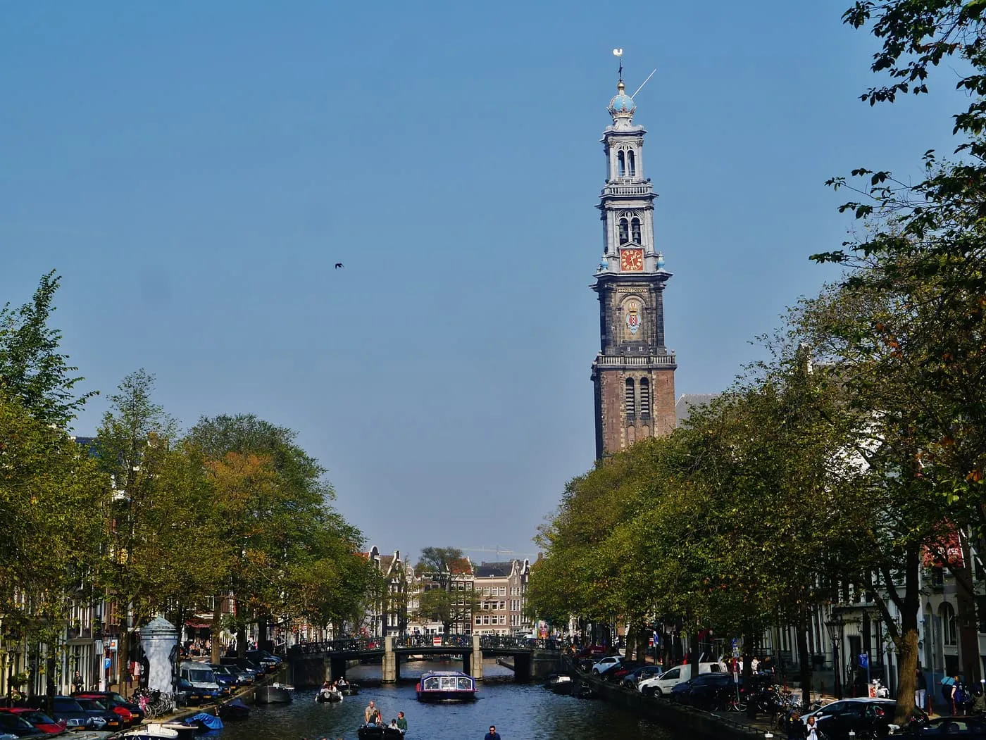 The Westerkerk tower rising above the trees along the Prinsengracht, with a tour boat and small boats on the canal under a bridge