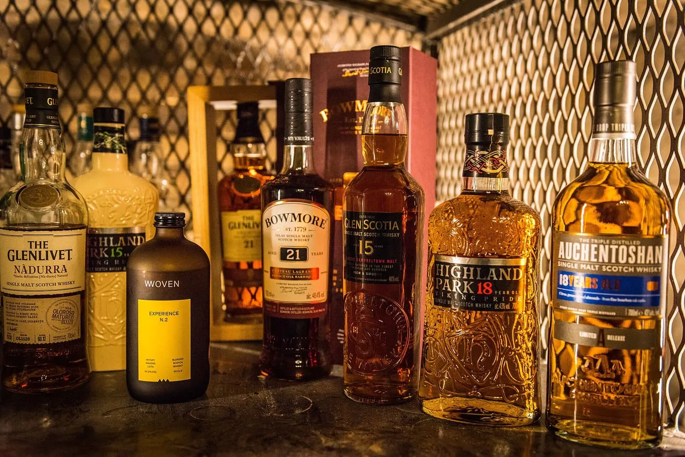 A row of Scotch whisky bottles, including Glenlivet, Bowmore, Glen Scotia, Highland Park and Auchentoshan, lit warmly on a shelf