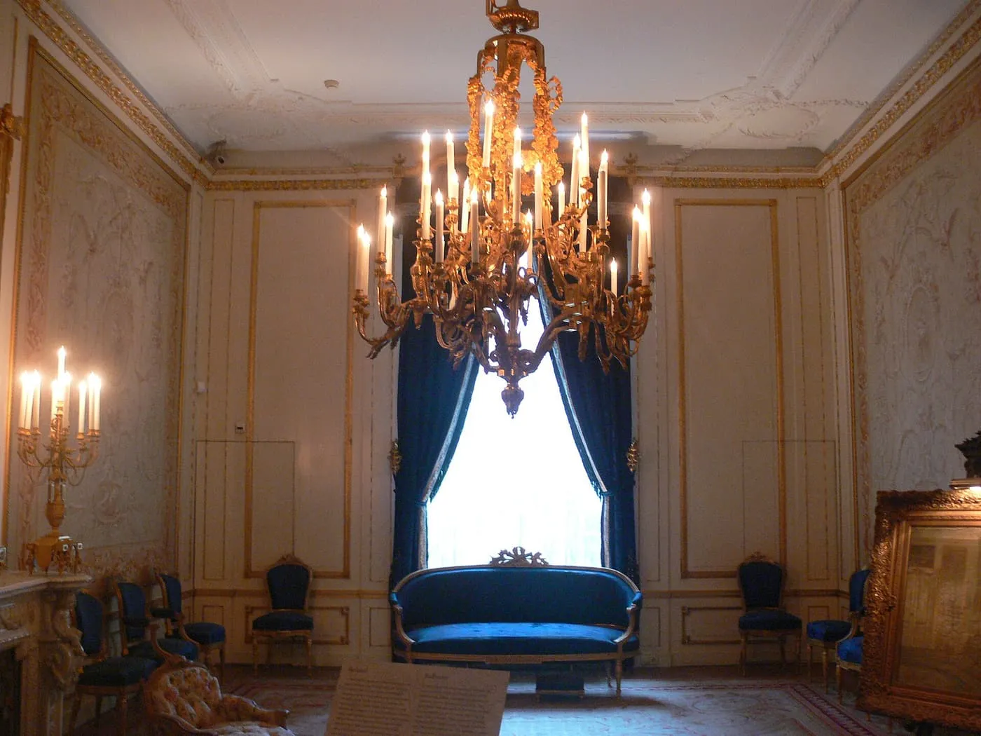A period room with a gilded chandelier of candles, a blue velvet sofa under a tall window and pale panelled walls