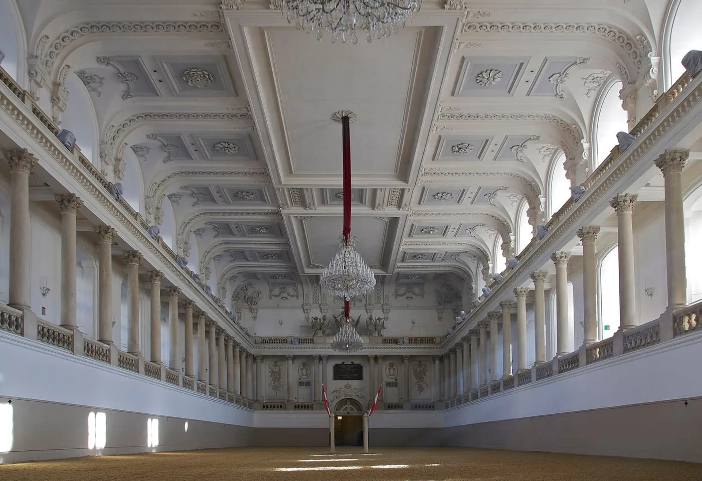 The empty Winter Riding School, a white hall with two tiers of columns, a chandelier and a sand floor