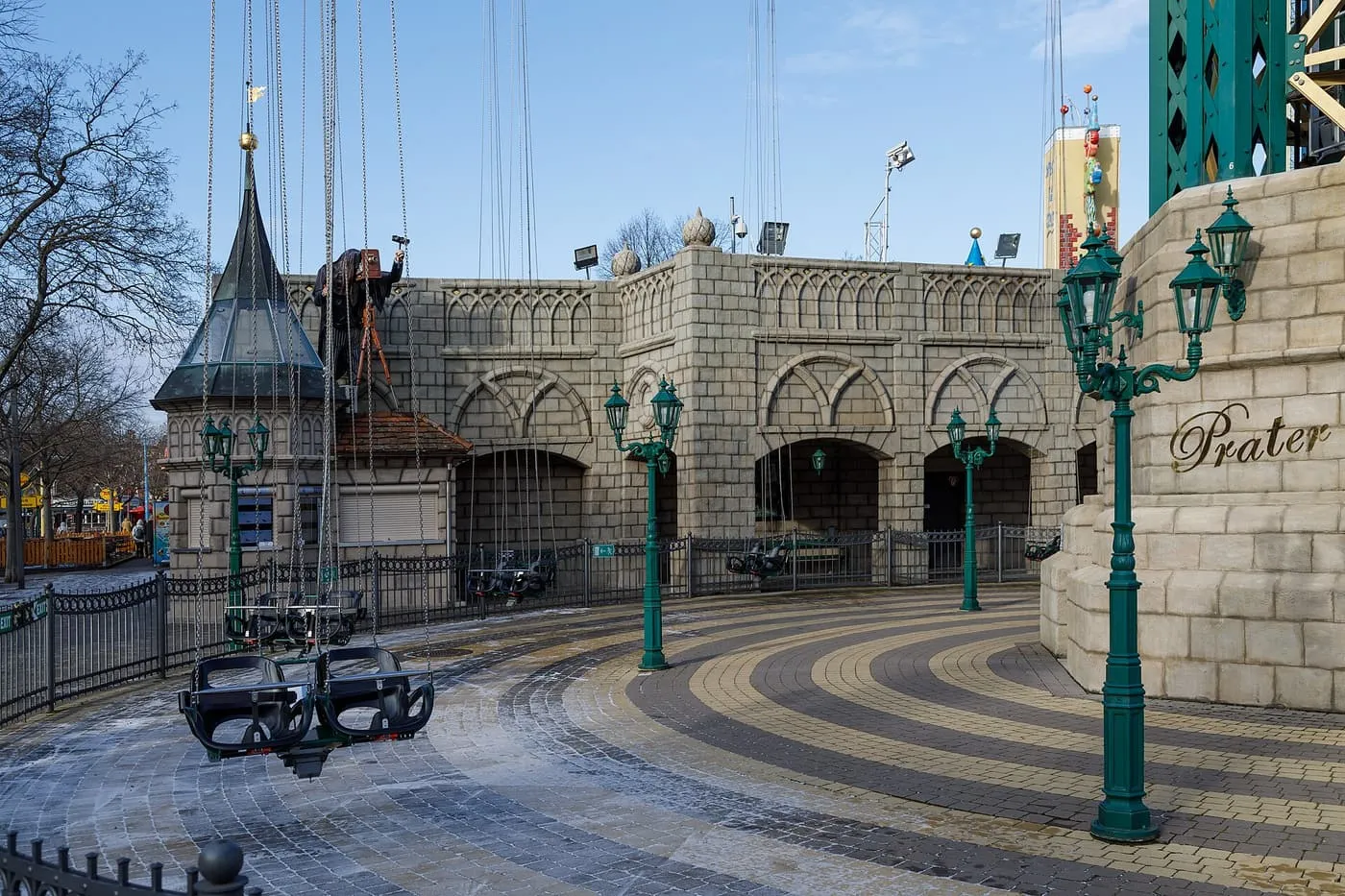 A chain-swing ride with empty seats hanging over a paved circle beside a stone wall and lamp posts, with Prater lettered on a pillar