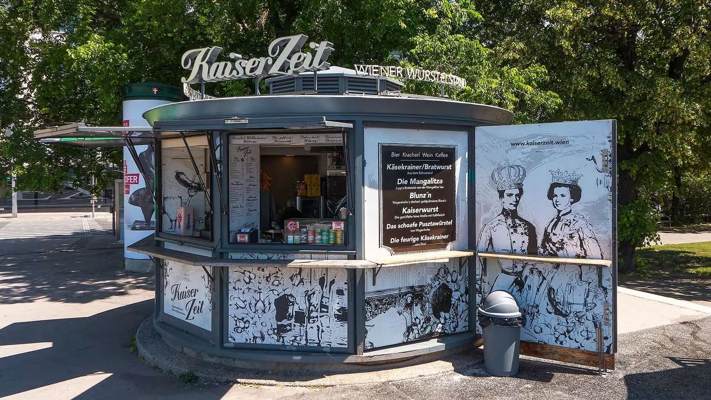 A round Würstelstand kiosk under trees, with a menu board of sausages on its side panel
