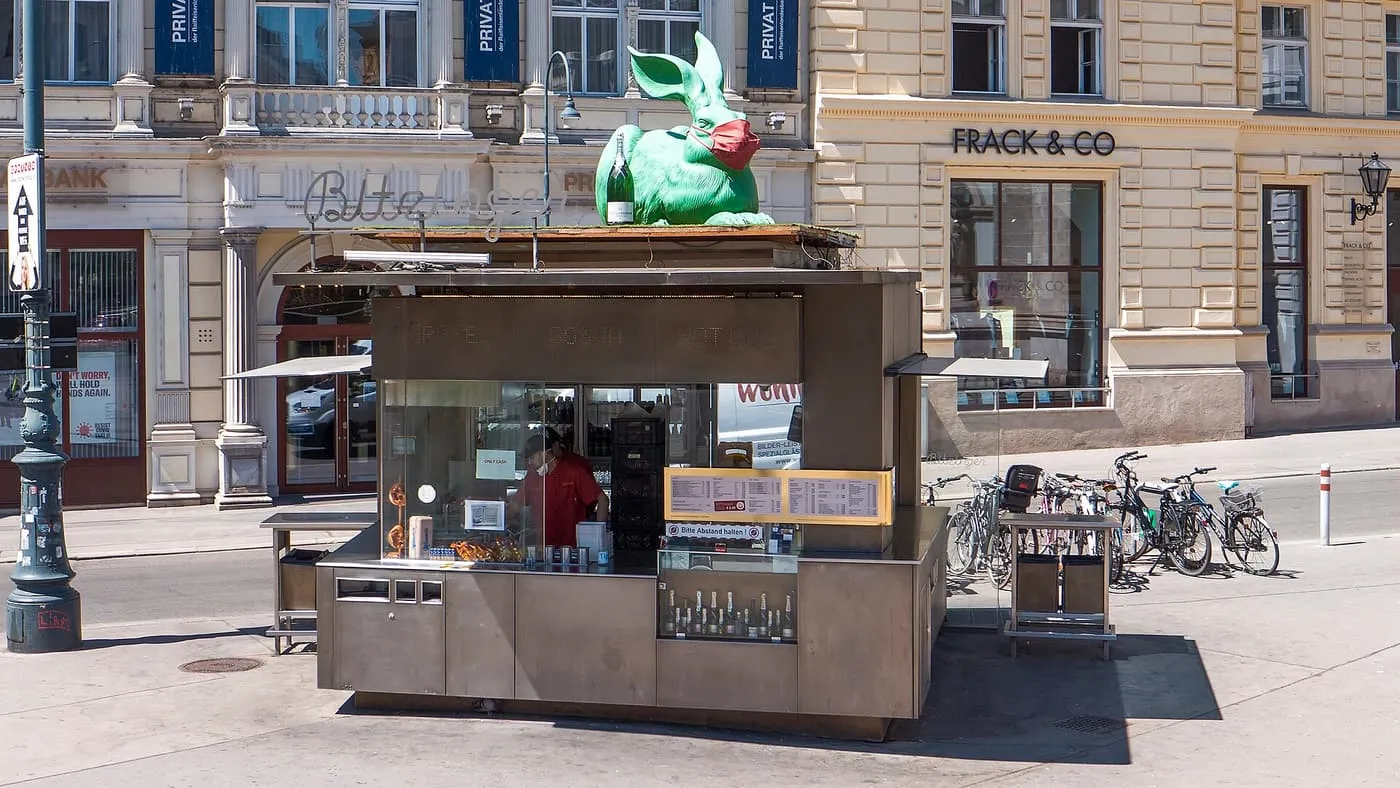 The Bitzinger Würstelstand, a steel kiosk with a green rabbit on its roof, on a square in front of cream-coloured buildings