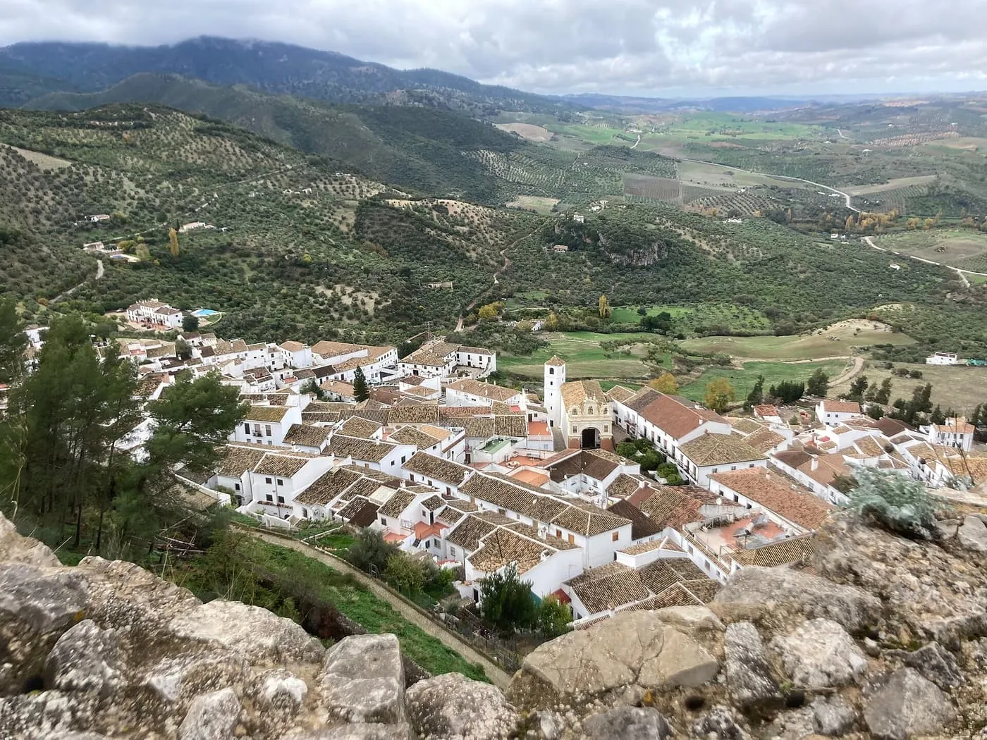 The white houses and terracotta roofs of Zahara de la Sierra seen from the rocks of the castle, with olive groves and mountains beyond