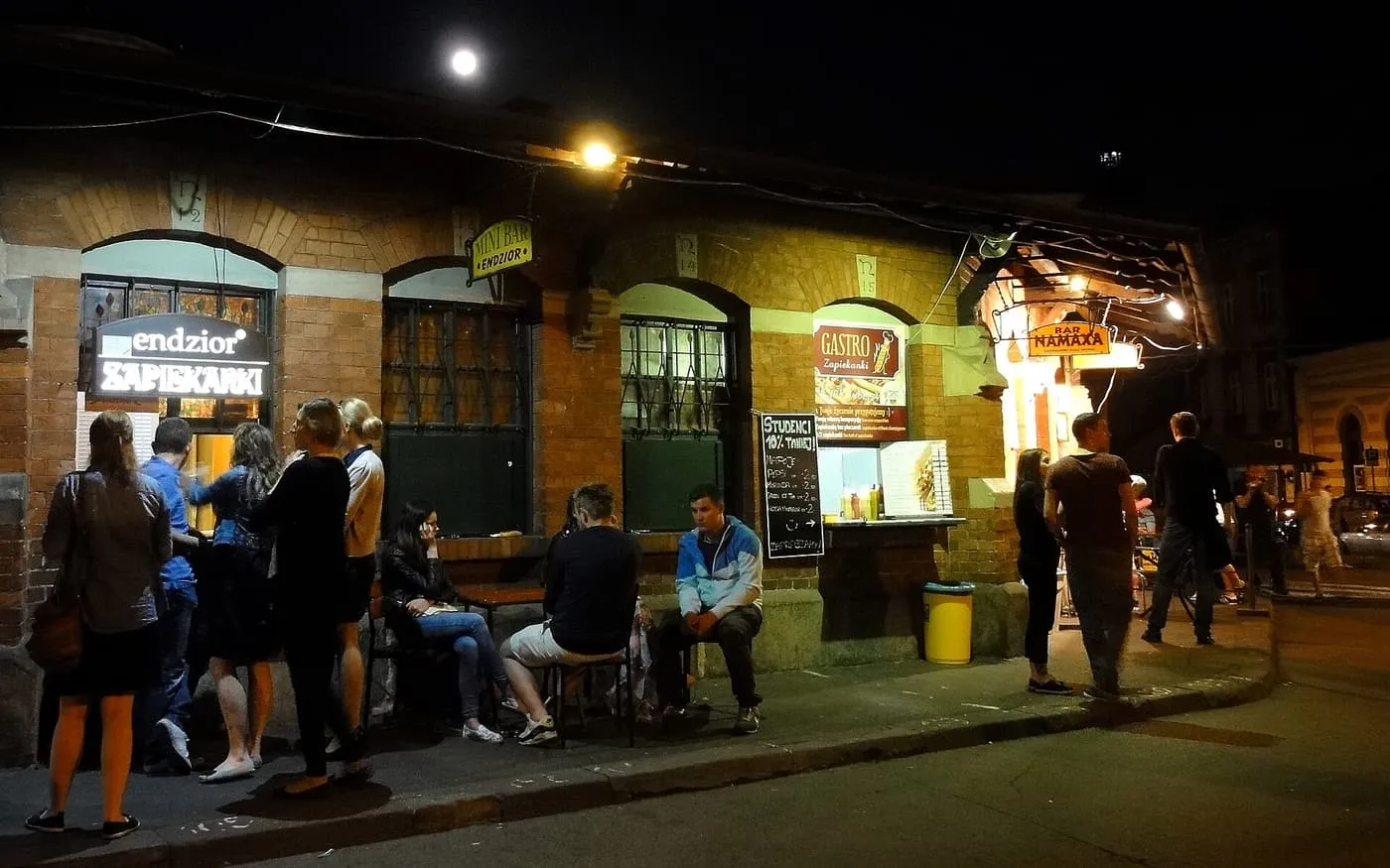 Zapiekanka stalls with lit signs in a red-brick building on Plac Nowy at night, with people queuing and sitting outside