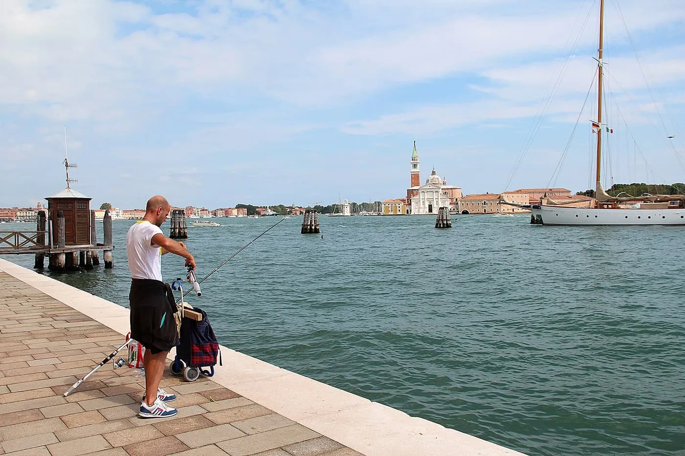 A man fishing from the paved Zattere quay, with San Giorgio Maggiore across the water and a sailing yacht beyond