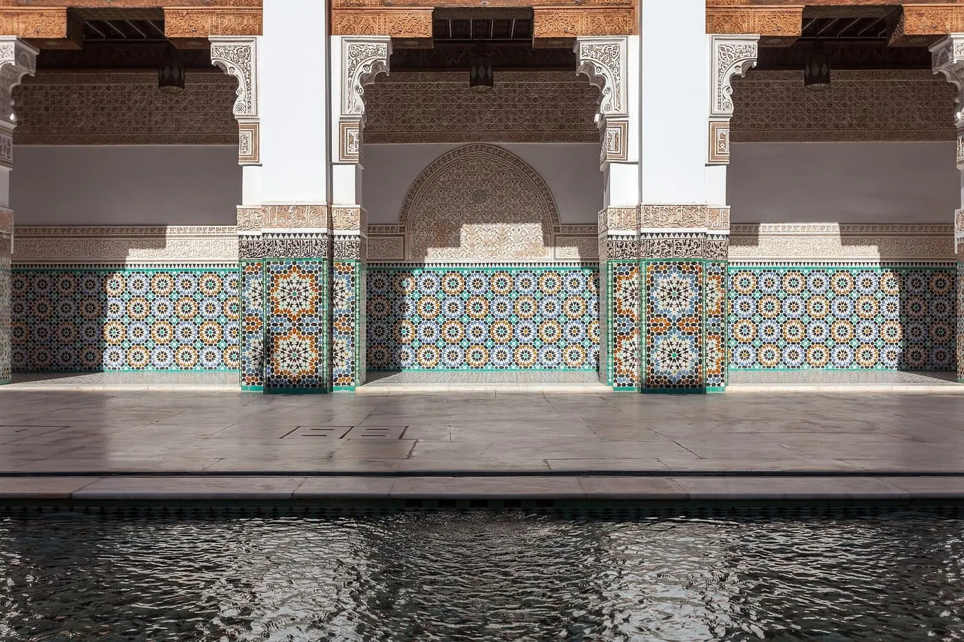 Zellige tilework and carved plaster arches on a gallery of the Ben Youssef Madrasa courtyard, above the edge of the pool